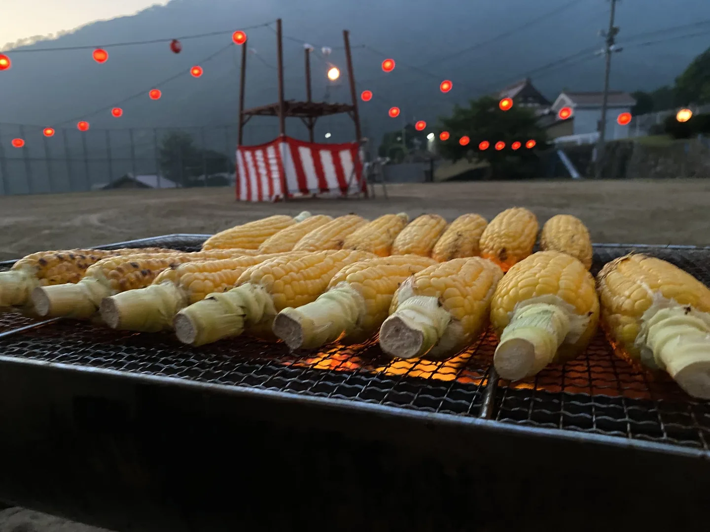 提灯とやぐらを背景に、網の上で焼かれるトウモロコシ