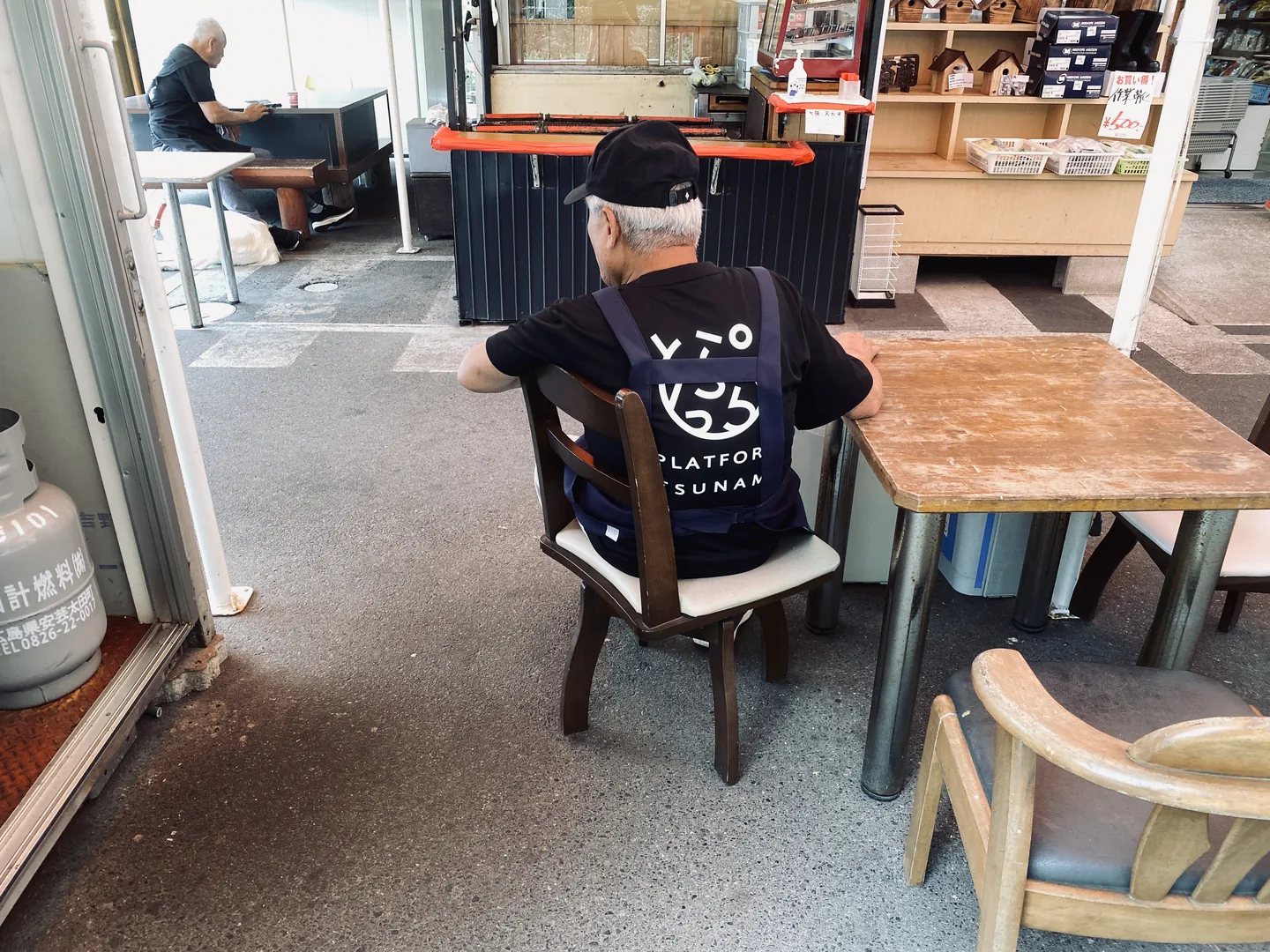 ロゴ入りTシャツにエプロンを重ね、施設内の椅子に座るスタッフ